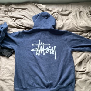 Mörkblå hoodie från Stüssy - Säljer en mörkblå hoodie från Stüssy med vit logga på bröstet och stor logga på ryggen. Tröjan har huva och en klassisk känguruficka framtill. Perfekt för en avslappnad streetwear-look.