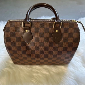 Damier speedy 25  handväska från Louis Vuitton - Säljer en klassisk brun handväska från Louis Vuitton damier 25 med rutigt mönster och guldiga detaljer. Väskan har två stadiga handtag, dragkedja och röd textilinsida. Kommer med lås och nyckel. Perfekt accessoar för dig som gillar lyxig stil.