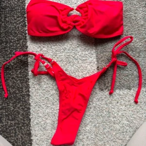 Röd bandeau bikini med knyt - Säljer en snygg röd bikini med bandeau-topp och knyt i sidorna på underdelen. Toppen har en unik ringdetalj i mitten och underdelen har smala band som knyts i sidorna, de är string. Perfekt för sommaren!