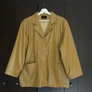 Beige skinnjacka från Kappa Erre - Säljer en beige skinnjacka från Kappa Erre Milano med klassisk krage och knäppning framtill. Jackan har två framfickor och en rak passform som ger en stilren look. Perfekt för dig som gillar tidlös design.