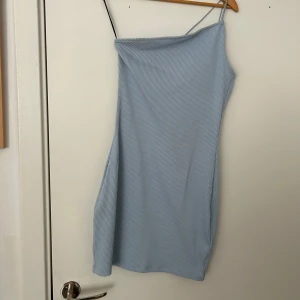 Ljusblå one shoulder klänning - Säljer en ljusblå kortklänning med smala axelband och one shoulder-design. Klänningen har en tight passform och är tillverkad i ett ribbat material som ger en snygg struktur. Perfekt för dig som gillar stilrena och trendiga plagg.