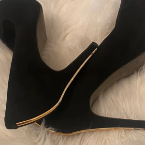 Svarta pumps med gulddetalj - Säljer ett par svarta pumps med hög klack och snygg guldfärgad detalj längs klacken och sulan. Skorna har rund tå och är tillverkade i mocka-liknande material. Perfekta för dig som vill sticka ut lite extra.
