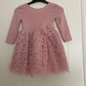 Rosa klänning med volangkjol och blomdetalj - Supersöt rosa barn klänning med långärmad överdel i mjuk bomull och fluffig kjol i tyll med volanger. Stor dekorativ blomma i tyll vid midjan. Perfekt för speciella tillfällen eller när man vill känna sig extra fin.