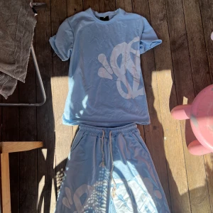 Ljusblå shorts och tröja  från syna world - Säljer ett par ljusblå shorts och tröja från syna world med vitt grafiskt tryck på ena benet. Shortsen har resår i midjan och snörning för extra komfort. Perfekta för sommardagar och har en avslappnad passform. Säljer det pågrund av att jag inte gillande passformen på dem 