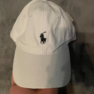 Vit keps från Polo Ralph Lauren - Snygg vit keps från Polo Ralph Lauren med klassisk broderad logga framtill och 'POLO' broderat baktill. Justerbart spänne i metall bak för perfekt passform. Perfekt accessoar för en stilren look.