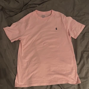 Ljusrosa t-shirt från Polo Ralph Lauren - Säljer en ljusrosa t-shirt från Polo Ralph Lauren med klassisk broderad logga på bröstet. T-shirten har rund halsringning och är tillverkad i mjuk bomull. Perfekt för en avslappnad och stilren look.