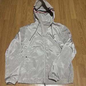 Moncler windbreaker  - Snygg windbreaker från Moncler med huva och dragsko. Jackan har dragkedja framtill, elastiska muddar och Moncler-logga på ärmen. Röd och svart rand på huvan ger en sportig touch. Perfekt för blåsiga dagar. (Priset är inte hugget i sten, hör av er vid minsta fundering)😁🤝