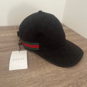 Svart keps från Gucci med grönt och rött band - Snygg svart keps från Gucci med diskret GG-mönster och klassiskt grönt och rött band på sidan. Justerbar rem baktill i svart läder med gulddetalj. Perfekt för dig som vill ha en stilren och lyxig accessoar.