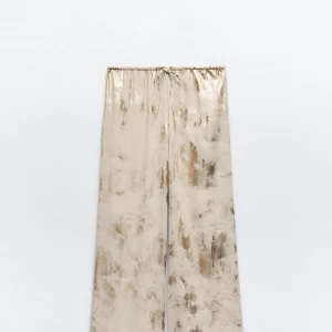 Beige och guldiga vida byxor från Zara - Säljer ett par beige vida byxor från Zara med skimrande guldmönster. Byxorna har resår i midjan med dragsko och är tillverkade i ett glansigt, mjukt material som ger en lyxig känsla. Perfekta för dig som vill sticka ut med stil.