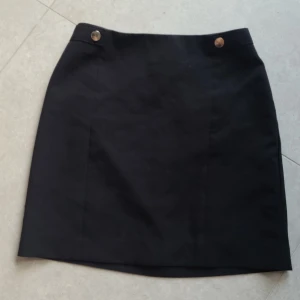 Svart kortkjol från H&M med knappar - Enkel svart kortkjol från H&M med två dekorativa metallknappar framtill vid linningen. Klassisk rak modell som är lätt att matcha. Perfekt basplagg i garderoben.