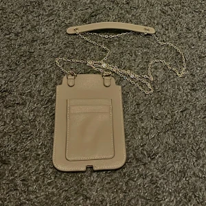 Oanvänd beige axelväska med justerbar kedja - En oanvänd beige axelväska i skinn med guldfärgad kedja och handtag. Väskan har ett fack på framsidan och en elegant metallogo på baksidan. Perfekt för mobilen, korten och nycklar! 💳🔑