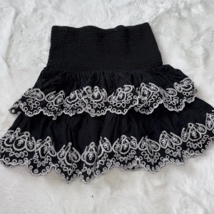 Svart kjol med broderade volanger från Lindex - Supersöt svart kjol från Lindex med bred smock i midjan och två lager volanger med vit brodyr. Kjolen har en kort modell och passar perfekt till en trendig look.