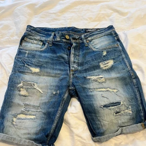 Blå jeansshorts med slitningar - Säljer ett par blå jeansshorts från Jack and Jones  med slitna detaljer och råa kanter. Klassisk femficksmodell med knappgylf och uppvikta benslut. Perfekta för en avslappnad stil.
