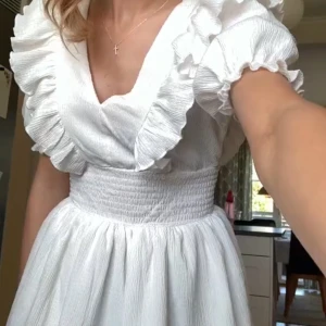 Vit volangklänning med v-ringning - Söt vit kortklänning med v-ringning, volangdetaljer och puffärmar. Smockad midja ger en markerad siluett och klänningen har dubbla lager i kjolen för extra volym. Perfekt för sommarens tillfällen. Endast använd en gång förra året🩷