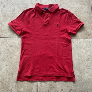 Röd pikétröja från Polo Ralph Lauren - Klassisk röd pikétröja från Polo Ralph Lauren med korta ärmar och krage. Tröjan har två knappar vid halsen och den ikoniska broderade loggan på bröstet. Perfekt för en stilren och avslappnad look.