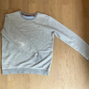 Ljusgrå sweatshirt från GANT - Säljer en stilren ljusgrå sweatshirt från GANT med broderad logga på bröstet. Tröjan har rund halsringning och långa ärmar med ribbade muddar. Perfekt för en avslappnad och klassisk look.