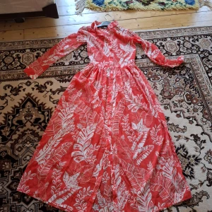 Röd långklänning med bladmönster - Säljer en röd långklänning med vita blad och växtmönster. Klänningen har lång ärm, markerad midja och knappar framtill. Perfekt för dig som gillar färgstarka och mönstrade plagg.