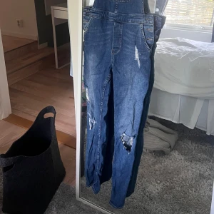 Blå denim hängselbyxor från H&M - Säljer ett par blå denim hängselbyxor från H&M med slitna detaljer på benen och klassiska fickor både fram och bak. Byxorna har raka ben och justerbara hängslen för en bekväm passform. Perfekt för en avslappnad och trendig look.