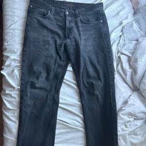 Svarta jeans från Nudie Jeans - Säljer ett par svarta jeans från Nudie Jeans med klassisk femficksdesign och silverfärgad knapp. Jeansen har en rak passform och är tillverkade i denim. Perfekta till vardags för en stilren look. Storlek 34/32 men passar även 36/32