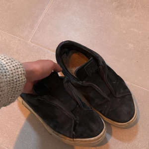 Svarta slip-on sneakers med vit sula - Svarta sneakers i slip-on modell med elastisk detalj över vristen och vit, platt sula. Skorna har en enkel och stilren design utan synliga loggor eller snörning. Passar perfekt till en avslappnad och urban stil.
