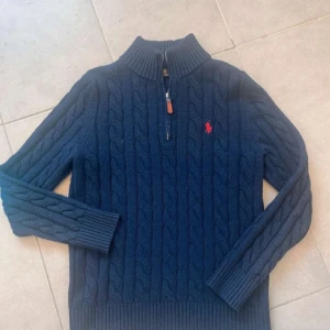 Blå kabelstickad tröja från Polo Ralph Lauren - Mörkblå kabelstickad tröja från Polo Ralph Lauren med halv dragkedja och hög krage. Tröjan har långa ärmar, ribbade muddar och röd broderad logga på bröstet. Perfekt för kyliga dagar.