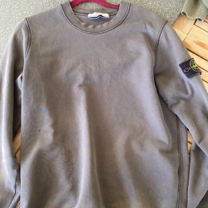 Grå sweatshirt från Stone Island - Säljer en grå långärmad sweatshirt från Stone Island med klassisk rund hals och logotyp-patch på vänster ärm. Tröjan har en enkel och stilren design, perfekt för en avslappnad look. Har använts en del gånger och har några defekter. Nypris 2500kr