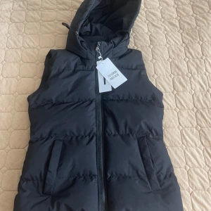 Svart dunväst med huva från Cherry Koko - Snygg svart dunväst från Cherry Koko med huva och dragkedja framtill. Västen har två fickor på sidorna. Aldrig använd. 