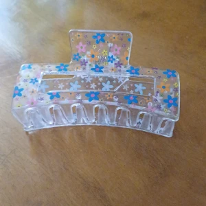 Blommig hårklämma i transparent plast - Säljer en söt hårklämma i transparent plast med färgglada blommor i blått, rosa, orange och vitt. Perfekt accessoar för att hålla håret på plats och ge en lekfull touch till din look. Har en liten spricka men går nog att limma