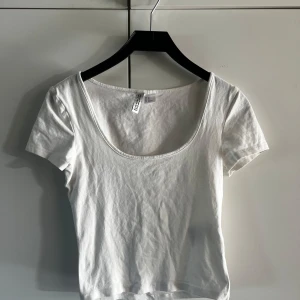 Vit croppad t-shirt med djup ringning från H&M - Säljer en enkel och stilren vit croppad t-shirt från H&M med korta ärmar och djup rund ringning. Perfekt basplagg att matcha med jeans eller kjol. Supermjuk och skön bomullskvalitet. Katt finns i hemmet! Använd sparsamt, kan till och med vara oanvänd!