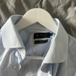 Ljusblå randig skjorta från Riley - Klassisk ljusblå skjorta från Riley med tunna vita och blåa ränder och knappar framtill. Skjortan har lång ärm och en stilren krage, perfekt för en clean och stilren look.