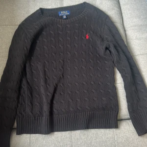 Svart kabelstickad tröja från Polo Ralph Lauren - Kabelstickad svart tröja från Polo Ralph Lauren med rund halsringning och lång ärm. Klassisk röd broderad logga på bröstet och ribbade muddar vid ärmslut och nederkant. Perfekt för en stilren och tidlös look.