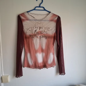 Mesh topp med tryck och långa ärmar - Säljer en transparent mesh topp med långa ärmar i vinröd och beige. Framsidan har ett stort tryck med texten 'LOVE YOU FOREVER' och motiv av hjärtan och band. Toppen har vågig kant vid ärmslut och nederkant.