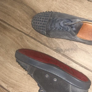 Svarta sneakers med nitar på tån - Säljer ett par svarta sneakers med rund tå och platt sula. Skorna har coola nitar framtill och är gjorda i mocka med svarta skosnören. Röd sula undertill ger en extra touch. Perfekta för dig som vill sticka ut!