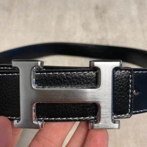 Svart skärp med H-spänne i silver - Säljer ett svart bälte från H3rmés med ett stort H-format spänne i silverfärgad metall. Bältet är gjort i präglat skinn med vita sömmar längs kanterna. Perfekt accessoar för att lyfta din outfit. Storlek 120cm. Priset ej hugget i sten 