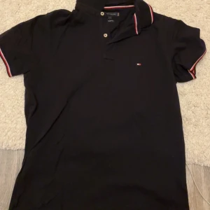 Mörkblå pikétröja från Tommy Hilfiger - Säljer en mörkblå pikétröja från Tommy Hilfiger med klassisk krage och knappar framtill. Tröjan har röda, vita och blå detaljer på krage och ärmslut samt liten logga på bröstet. Tillverkad i ekologisk bomull och har en slim fit passform. Den är storlek L men liten i storleken. Använd 1-2 gånger. Perfekt skick. Ordinarie pris ca 800 kr.