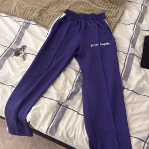 Lila träningsbyxor från Palm Angels - Säljer ett par lila träningsbyxor från Palm Angels med vita sidoränder och elastisk midja. Byxorna har raka ben och fickor på sidorna. Perfekta för en sportig look. 6/10 skick