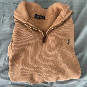Beige halvzip tröja från Polo Ralph Lauren - Säljer en beige långärmad halvzip-tröja från Polo Ralph Lauren. Tröjan har en klassisk krage med dragkedja och den ikoniska broderade loggan på bröstet. Perfekt för lager-på-lager och stilren vardagslook. Skriv vid minsta lilla fundering!