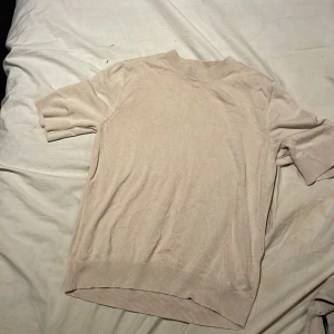 Beige kortärmad stickad tröja - Säljer en beige stickad tröja med korta ärmar och ribbade muddar vid hals, ärmslut och nederkant. Perfekt för lager-på-lager och enkel att matcha med olika outfits.