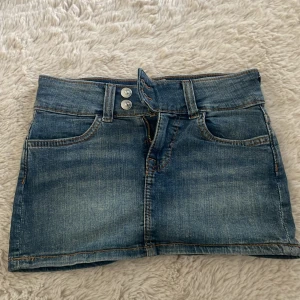 Jeanskjol från H&M Divided - Säljer en klassisk blå jeanskjol från H&M Divided med två knappar framtill och femficksmodell. Kjolen är kort och har en rak passform, perfekt till sommaren. Snygga detaljer med dubbla sömmar och slitningar. XXS/XS