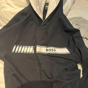 Svart zip-hoodie från BOSS - Svart hoodie med dragkedja från BOSS. Har vit rand och grafiskt tryck över bröstet samt snörning i huvan. Insidan av huvan är ljusgrå. Perfekt för en avslappnad stil.