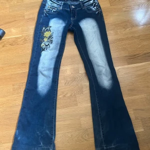 Blå jeansbyxor med broderad detalj från Crazy Age - Unika blå jeansbyxor med broderade och glittriga detaljer i gult och svart på både fram- och bakfickor. Byxorna har bootcut-modell och tydliga slitningar framtill för en cool look. Perfekta för dig som vill sticka ut.