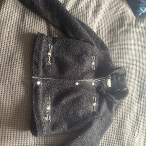 Svart teddyjacka från Holly & Whyte - Använd 2 ggr. Mysig och dressad stilren svart teddyjacka från Holly & Whyte med dekorativa knappar och fickor framtill. Jackan har lång ärm och en klassisk, lite boxig passform. Perfekt för kyligare dagar och enkel att styla till olika outfits.