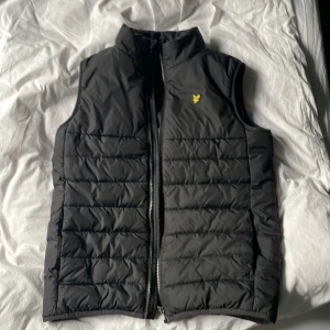 Svart dunväst från Lyle & Scott Junior - Snygg svart dunväst från Lyle & Scott Junior med gul logga på bröstet. Västen har ståkrage, quiltad design och dragkedja framtill. Perfekt för lager-på-lager under kyliga dagar.