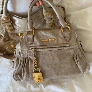 Miu Miu Vitello Leather Bag - Snyggaste Miu Miu Vitello handväskan! Supersnygg beige färg med gulddetaljer. Äkta läder. Extra touch med ett lås på framsidan och flera fack med fickor. Långt band tillkommer även. Fint skick, men finns tecken på användning (se alla bilder). Snyggaste väskan som ger liv till vilken outfit som helst! Köpt second hand så kvitto finns ej, men köp gärna via Vinteds artikelverifiering! Går att köpa via köp nu eller hämtas hemma hos mig i Nacka 🤍 
 Mått:  Bredd: 30 cm Höjd: 20 cm Djup: 12 cm
