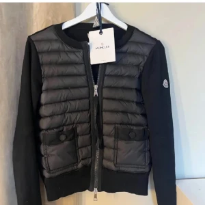 Moncler cardigan  - Säljer en svart pufferjacka från Moncler med quiltad framsida och två fickor med knappar. Jackan har rund halsringning, långa ärmar och dragkedja framtill. I nyskick, taggarna följer med och det finns qr kod. Skicka prisförslag så accepterar jag köpet via plick!