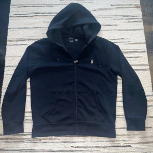 Mörkblå hoodie med dragkedja från Polo Ralph Lauren - Säljer en mörkblå hoodie från Polo Ralph Lauren i storlek M. Tröjan har dragkedja framtill, huva med snörning och klassisk broderad logga på bröstet. Praktiska fickor framtill och mjukt material som passar perfekt till vardags. OBS har lite noppor på sig men inte trasig någonstans! Köpt från NK för 2000kr