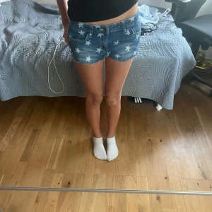 Jeansshorts med stjärnmönster - Säljer ett par blå jeansshorts från Lindex  med vita stjärnor över hela tyget. Shortsen har råa kanter, fem fickor och knappgylf. Perfekta för sommaren och ger en cool, avslappnad look.