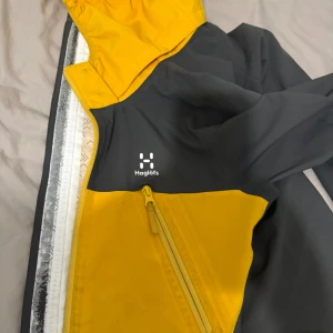 Gul och mörkgrå vindjacka från Haglöfs - Säljer en snygg vindjacka från Haglöfs i gul och mörkgrå färg. Jackan har huva, dragkedja framtill och praktiska fickor. Perfekt för utomhusaktiviteter och har en sportig look. Materialet är syntet och jackan är långärmad.