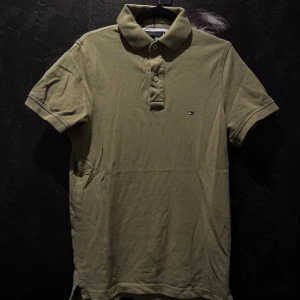 Olivgrön pikétröja från Tommy Hilfiger - Snygg olivgrön pikétröja från Tommy Hilfiger med klassisk krage och knappar framtill. Liten broderad logga på bröstet. Perfekt för en stilren och avslappnad look.