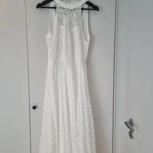 Vit långklänning med spets och knyt i nacken - Vit långklänning med vacker spets och blommigt mönster. Klänningen är ärmlös och har hög halsringning samt knyt i nacken. Perfekt för sommarens speciella tillfällen.